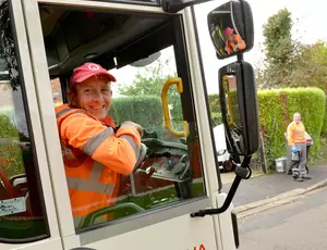 Photo showing a cheerful driver in a bin collection truck, with another crew member in the backgroun collecting bins.