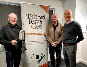 Photo of a volunteer holding a kettle and screw driver, beside a Telford Repair Cafe pull up banner, and representatives from Veolia and Telford & Wrekin Council