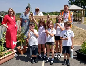 Photo shows a group of five children holding vegetables, with three adults, centred around a vegetable selling stand in a gardening area on a sunny day.