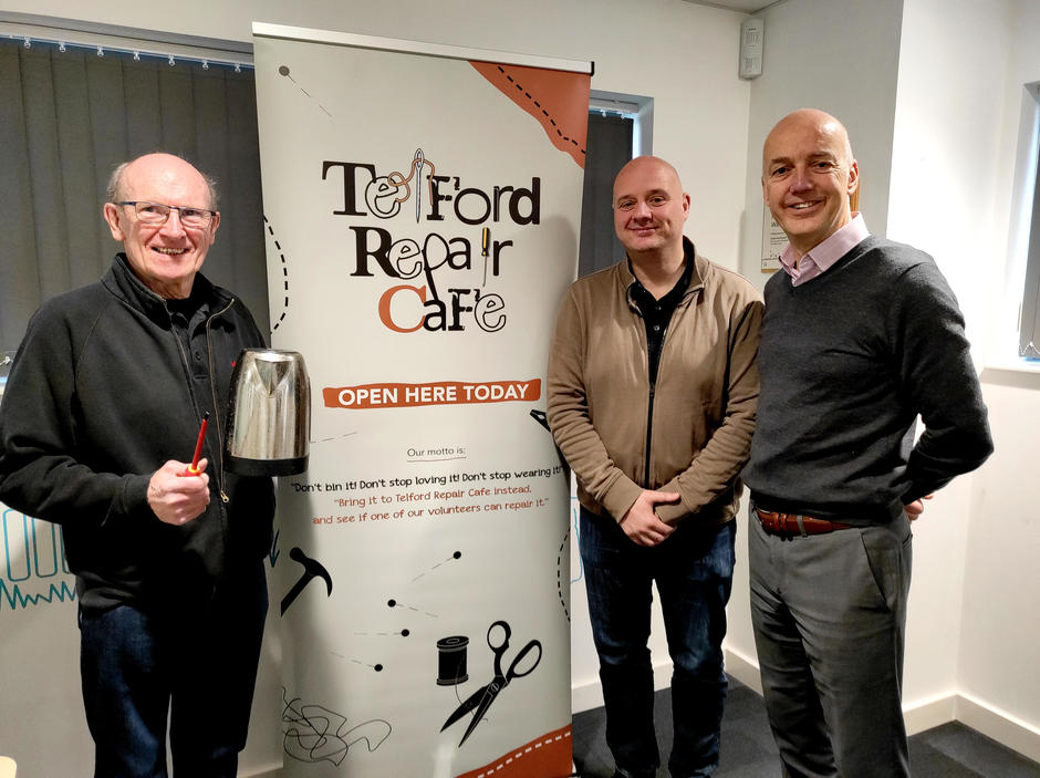 Photo of a volunteer holding a kettle and screw driver, beside a Telford Repair Cafe pull up banner, and representatives from Veolia and Telford & Wrekin Council