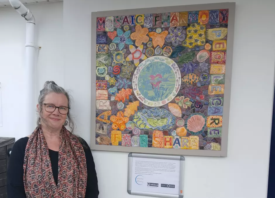 Photo shows a large and multicoloured artwork with individual tiles depicting leaves, flowers and the words People Earth Care and Fare Share. With Rosalyn Donati, Creative Lead